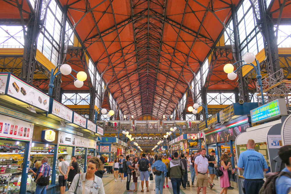 View down the middle of the inside of the Grand Market in Budapest, Hungary. Food stalls either side and lots of people walking.