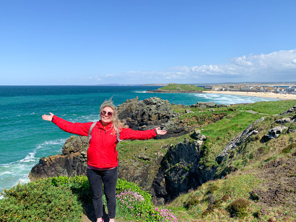 Me in a red jacket standing with arms open wide with a view of St Ives Bay in the background