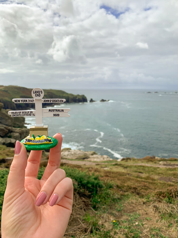 My hand holding a miniature sign of Land's End with a view of Land's End behind it