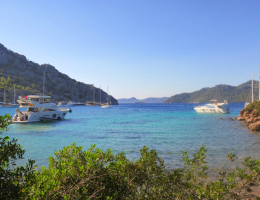 Turquoise blue sea, with mountains stretching into the distance, shrubs in the foreground and a few boats and yachts anchored in the water.