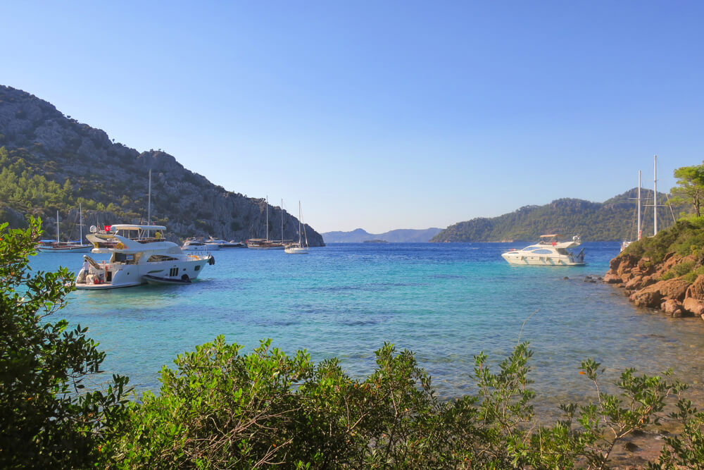 Turquoise blue sea, with mountains stretching into the distance, shrubs in the foreground and a few boats and yachts anchored in the water.