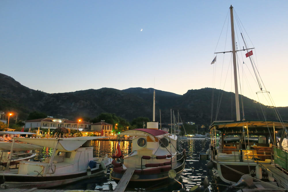 Three boats in the water, with Selimiye's promenade and mountains in the background and a crescent moon in the sky.