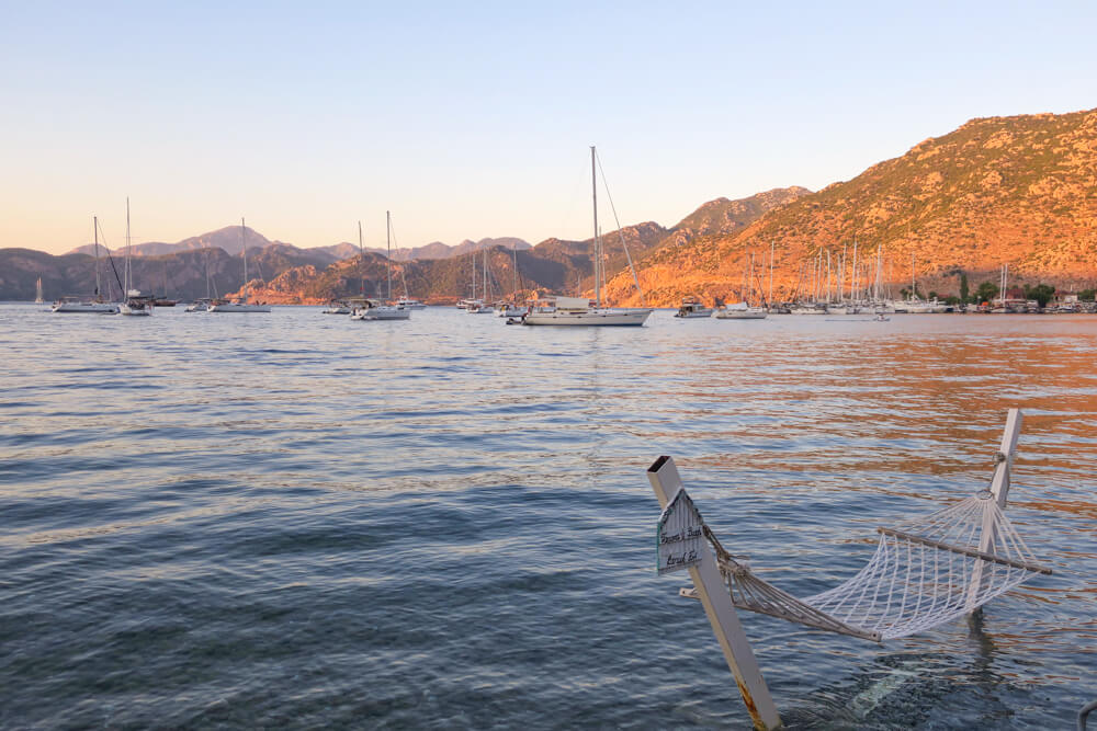 A string hammock in the water at Selimiye, with yachts anchored and mountains lit up by the hour before sunset in the background.