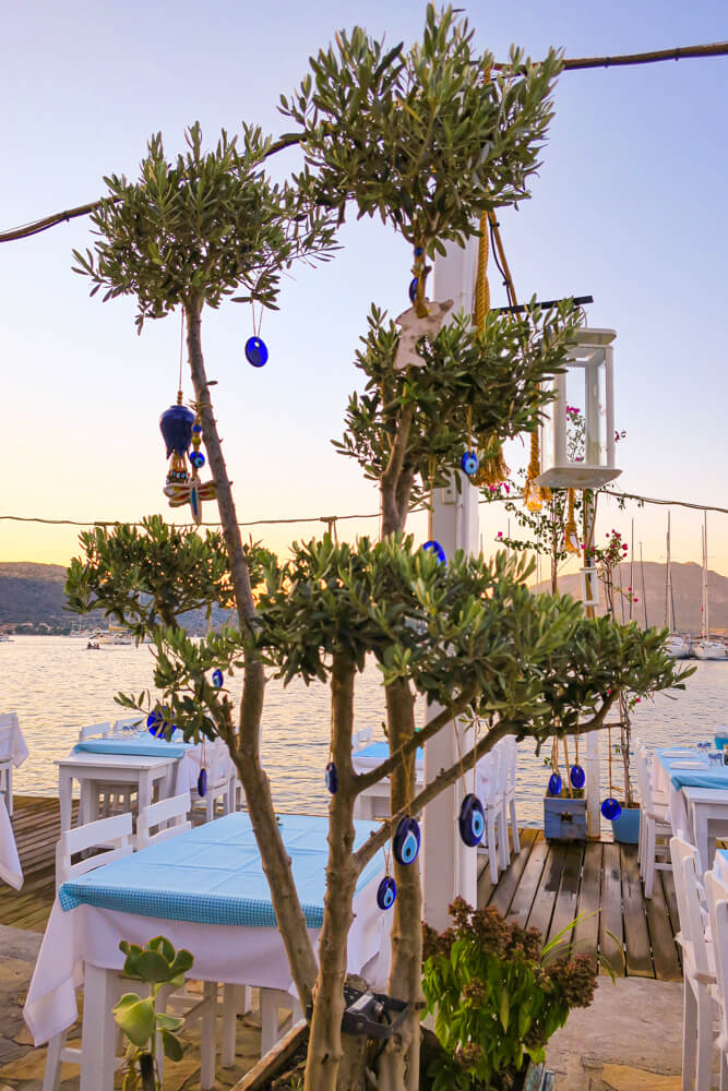 An olive tree in Selimiye decorated with hanging blue Turkish evil eyes of varying sizes, with restaurant tables, the sea and mountains behind.