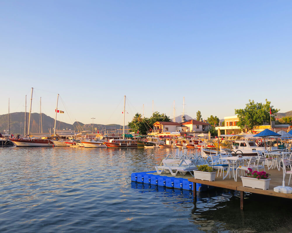 View of Selimiye harbour at golden hour, with the sea in the foreground and the boats and mountains in the background. A restaurant is on the right hand side.