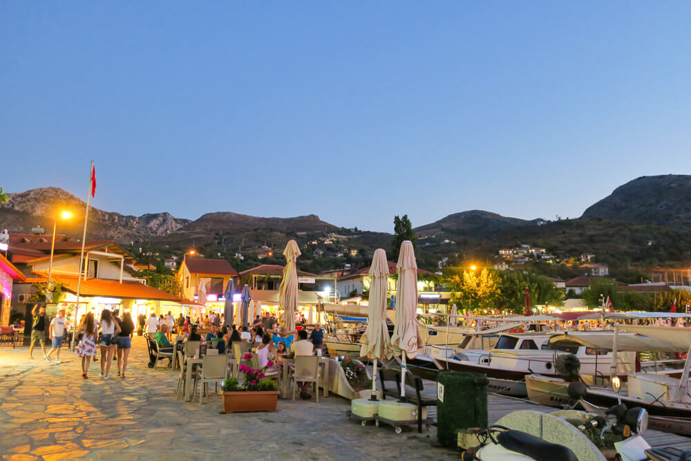 A view of part of the promenade around Selimiye harbour in the evening, with boats, restaurant tables, lights and mountains.