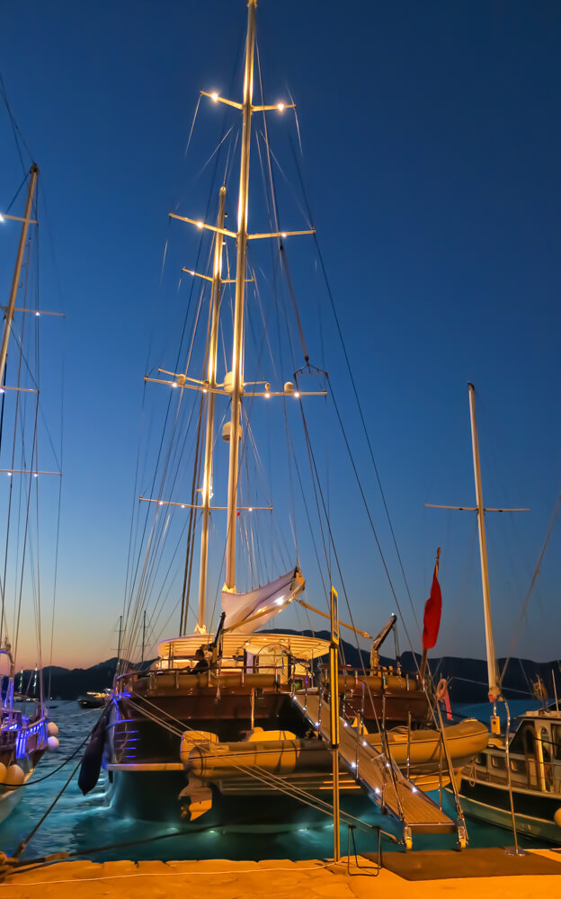 A tall, expensive yacht in Selimiye, lit up at night with lighting under the boat, in the water and up the masts. Mountains are in the background.