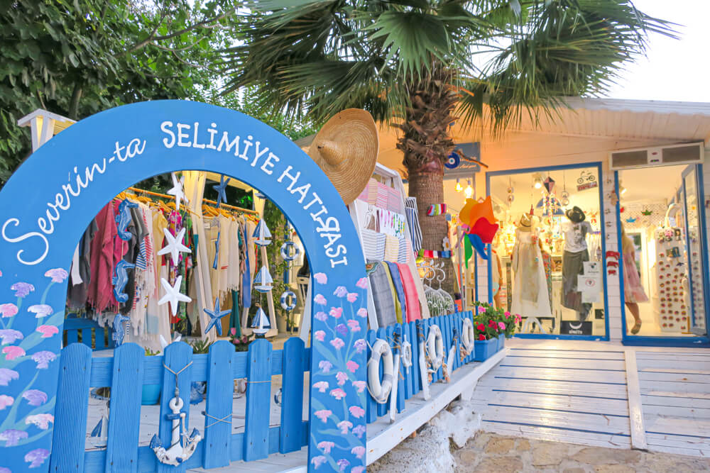 A blue fence with an arched shop sign, leading to a souvenir shop with a palm tree outside and lots of clothing on racks.