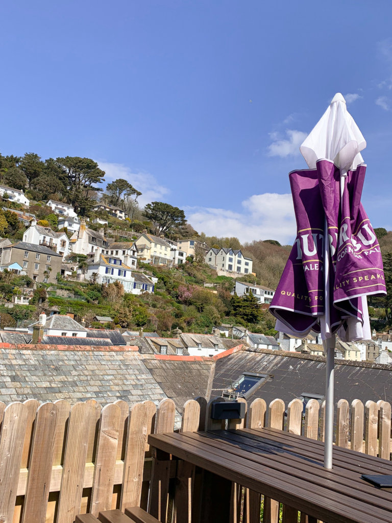 A closed purple umbrella sticks out of a picnic bench table on the rooftop of the Three Pilchards pub in Polperro on the south coast of Cornwall, with a tree and house filled hillside in the background.