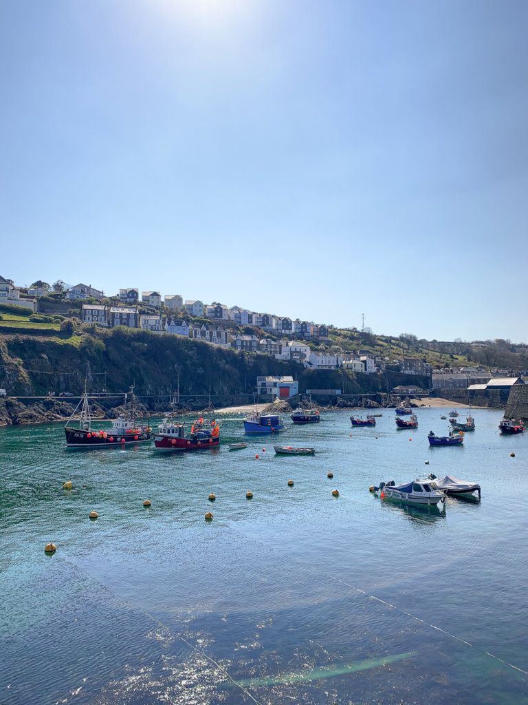 Fishing boats bob in the turquoise water of Mevagissey harbour on the south coast of Cornwall, with terraced houses diagonally spreading up the hillside in the background.