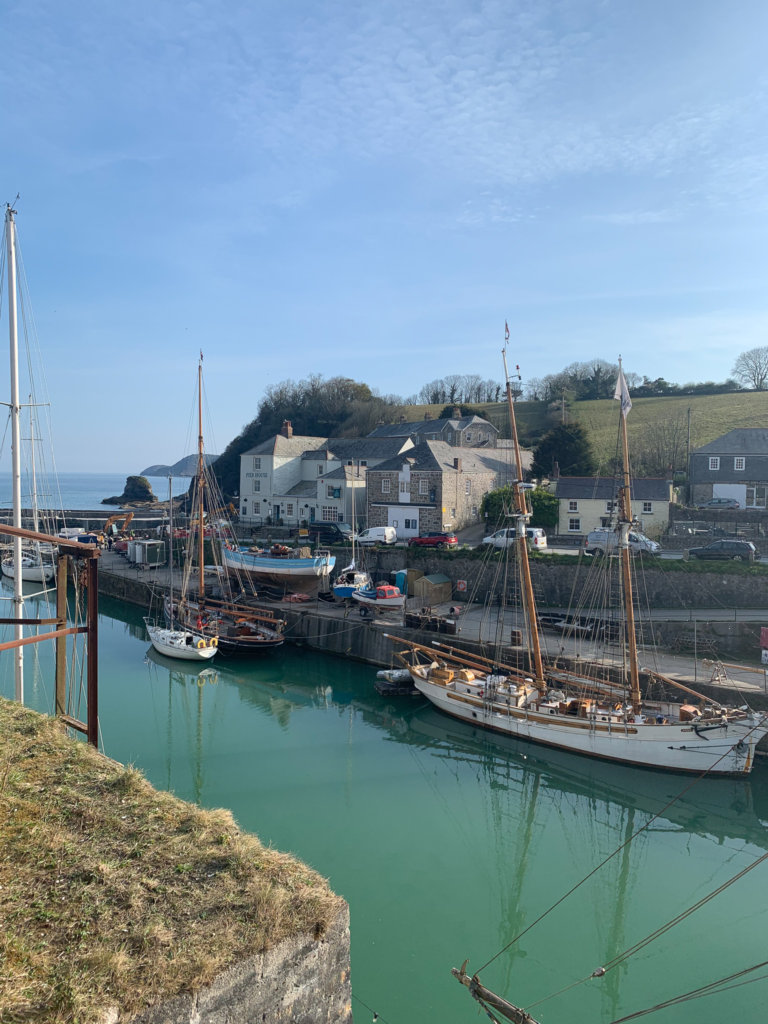 Tall wooden ships sit in the calm turquoise water of Charlestown port on the south coast of Cornwall, with cottages behind and the sea in the distance.