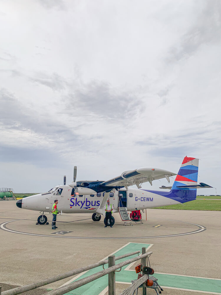 A 19-seater Skybus plane sits on the tarmac ready for passengers to board to fly to the Isles of Scilly. The plane is white and the tail is blue and red. Blog post - Top Tips for the Isles of Scilly.