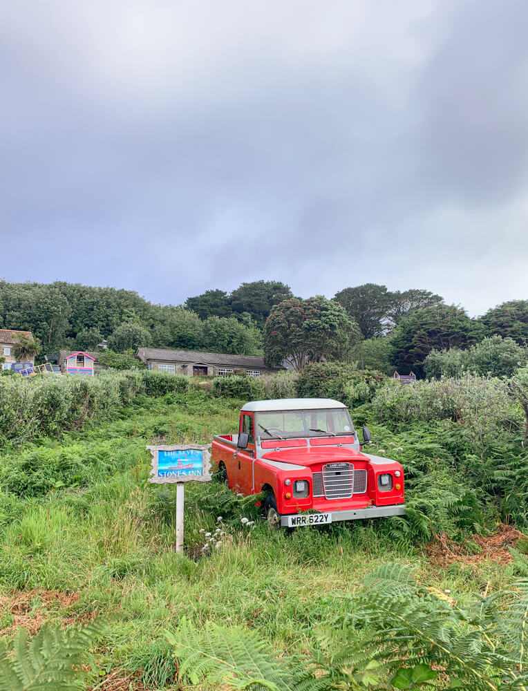 An old red truck sits in the middle of an overgrown field, with a blue sign to its left that says "The Seven Stones Inn". Blog post - Top Tips for the Isles of Scilly.