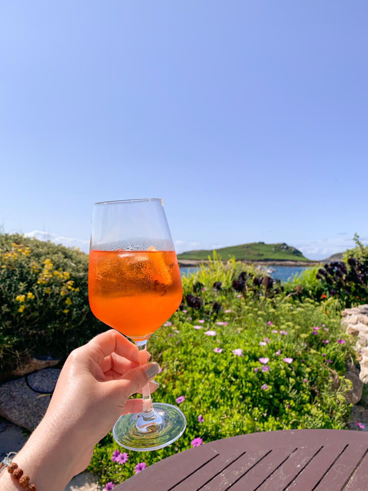 Nina holds a glass of Aperol Spritz into the air, with green plants, flowers and a peek of the ocean in the background. Blog post - Top Tips for the Isles of Scilly.