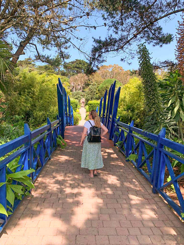 Nina walks away from the camera in the centre of a bright blue wooden bridge, with trees surrounding the bridge on all sides. Blog post - Top Tips for the Isles of Scilly.