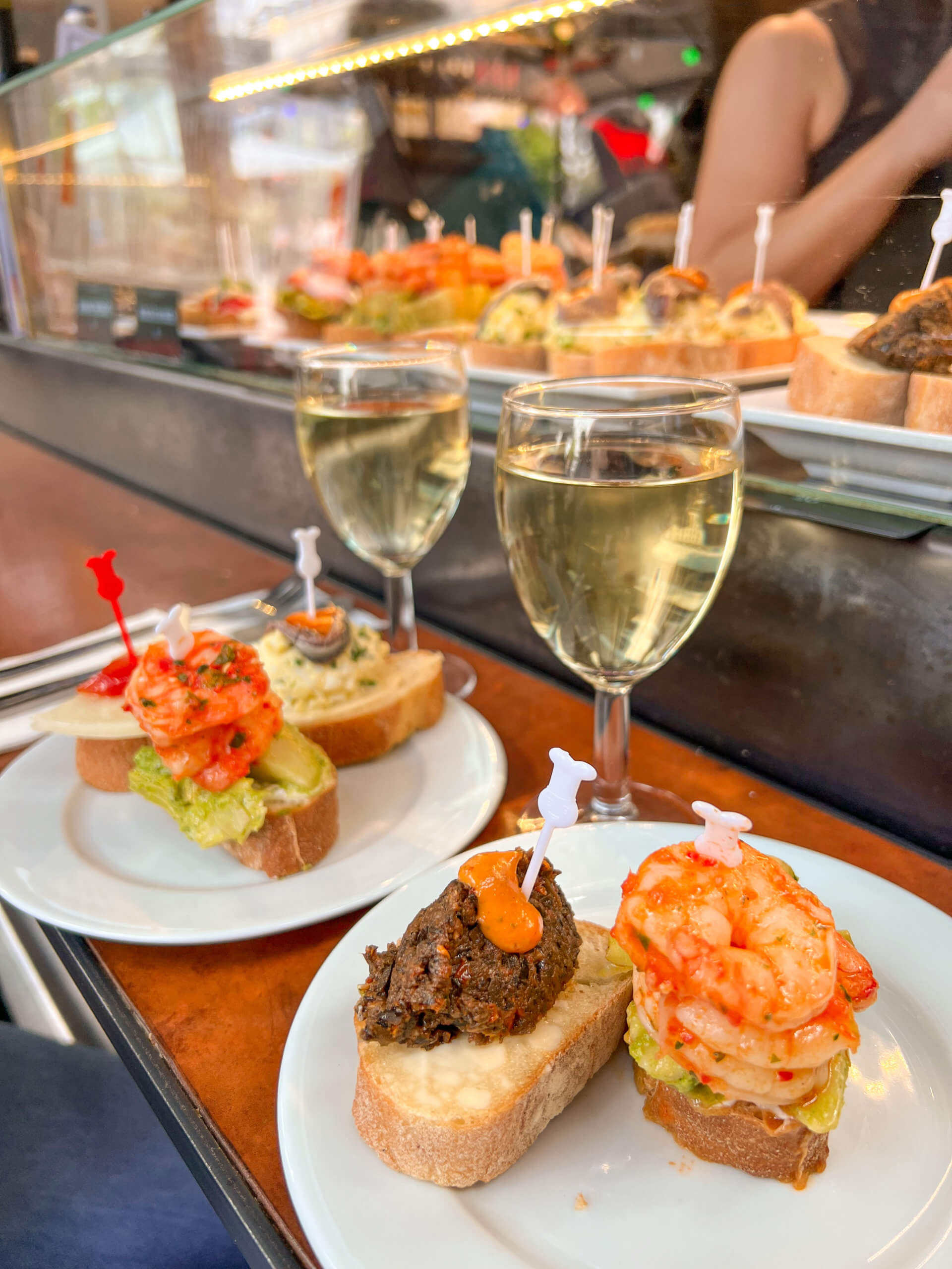 Two plates of tapas sit in front of two glasses of white wine, with more tapas enclosed in a glass counter behind. One of my top 10 places to visit in Copenhagen.