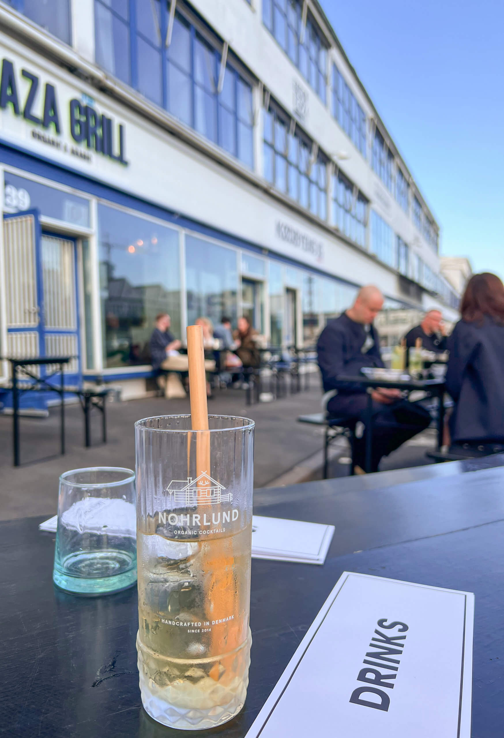 A drink with bamboo straw is in the foreground on a table, with a menu next to it saying "drinks". Picnic benches are behind with the white and blue buildings of the meat packing district. One of my top 10 places to visit in Copenhagen.