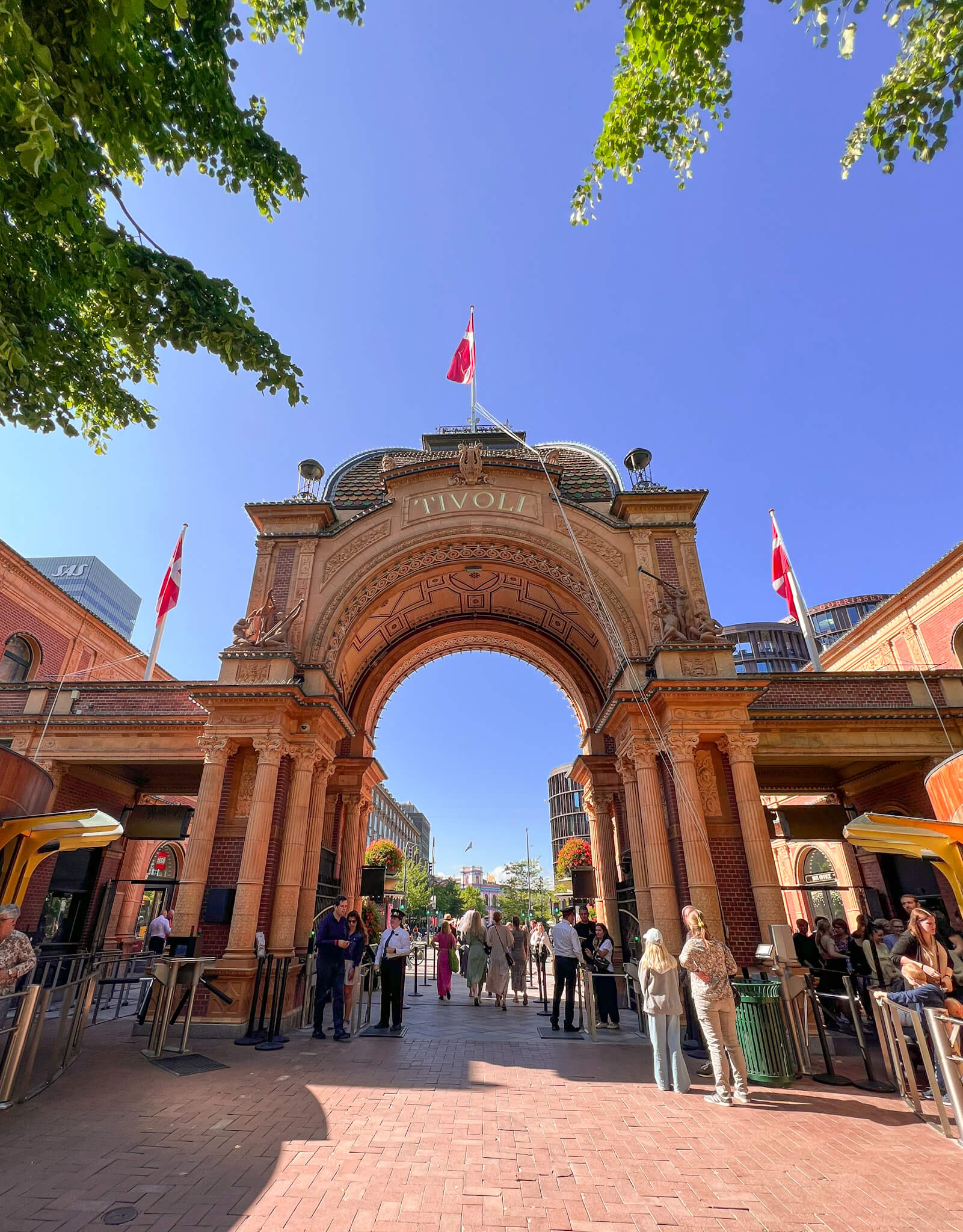 View of the inside stone arch gates of Tivoli Gardens, with people queuing to get in and the Danish flag flying above. One of my top 10 places to visit in Copenhagen.