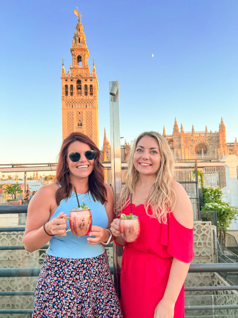 Two women stand holding cocktails in front of the Seville Cathedral on a rooftop bar - 10 things to do in Seville