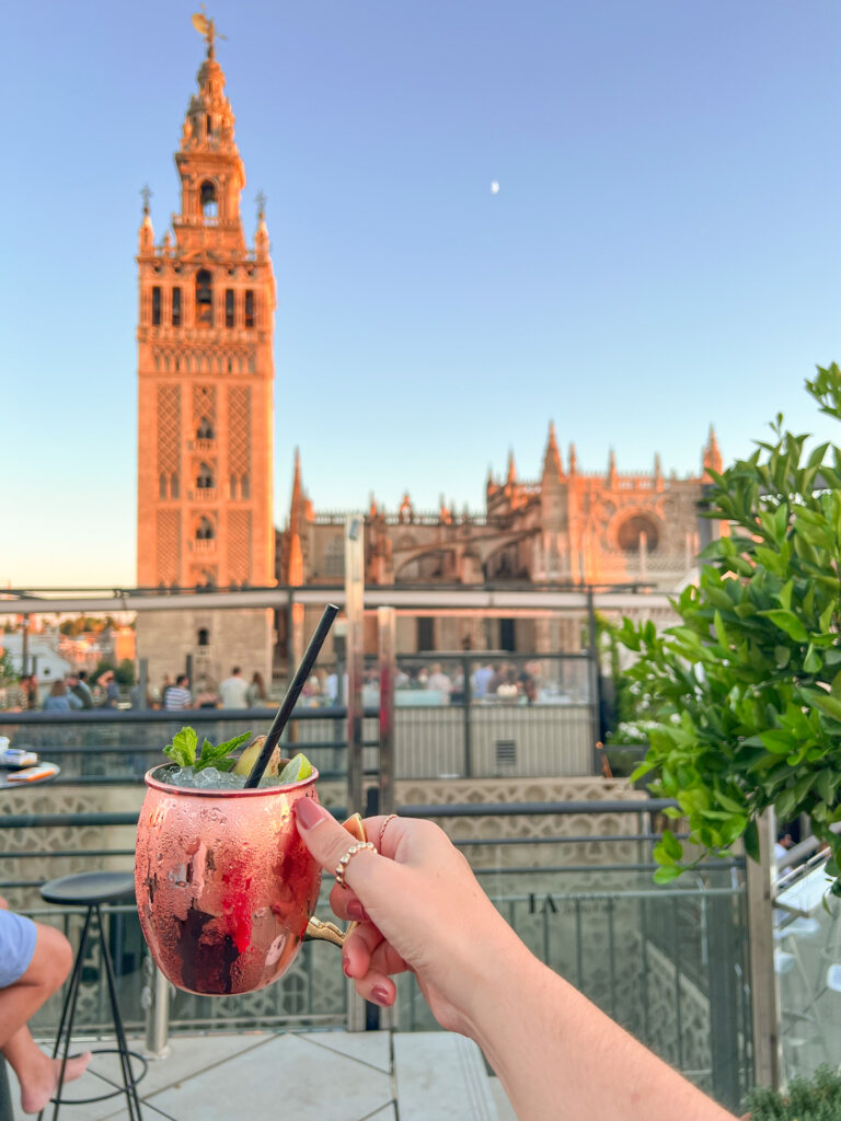 Hand holds out a Moscow Mule cocktail with a backdrop of the Seville cathedral - 10 things to do in Seville